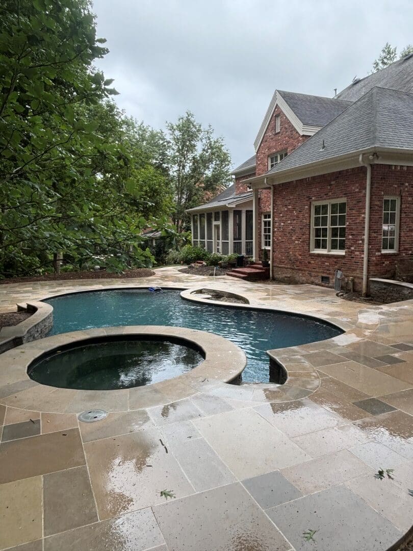 Freeform pool and spa with a French-pattern limestone deck behind a brick home